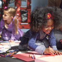 Cheerful Girl Drawing On Paper While Sitting By Teacher At Preschool